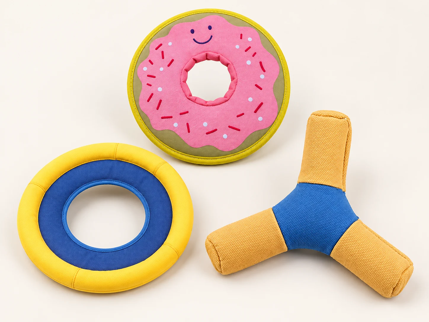Three fabric-covered dog fetch toys: pink doughnut-shaped ring, yellow-and-blue circular flying ring and orange-and-blue three-arm design