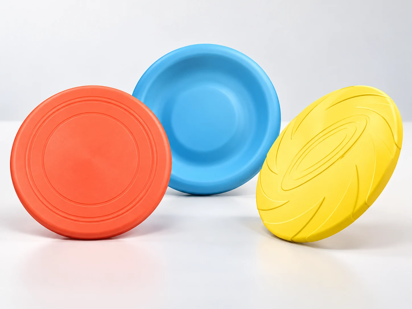 Three round dog training flying discs in coral, blue and yellow, shown at different angles to reveal their shallow profiles and rim shapes