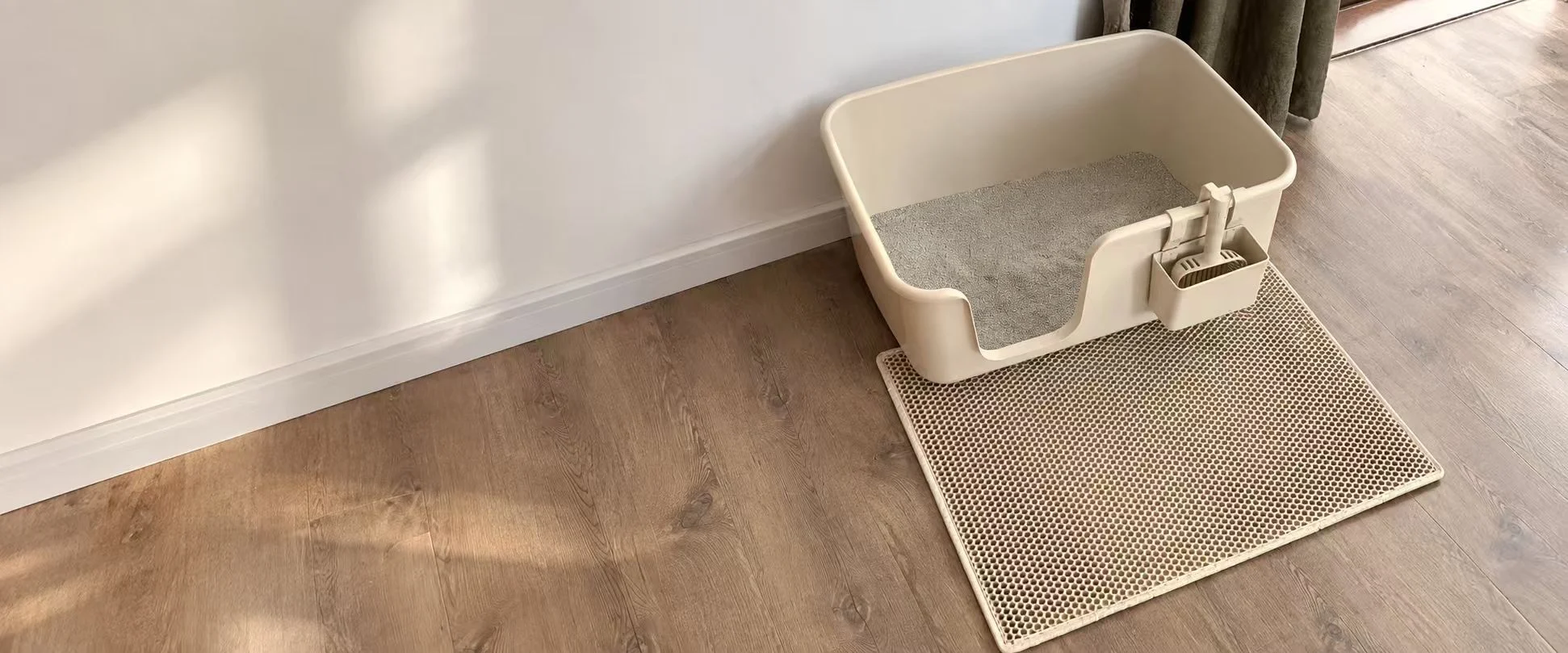 Beige open cat litter tray with a scoop and litter-catching mat on a wooden floor.