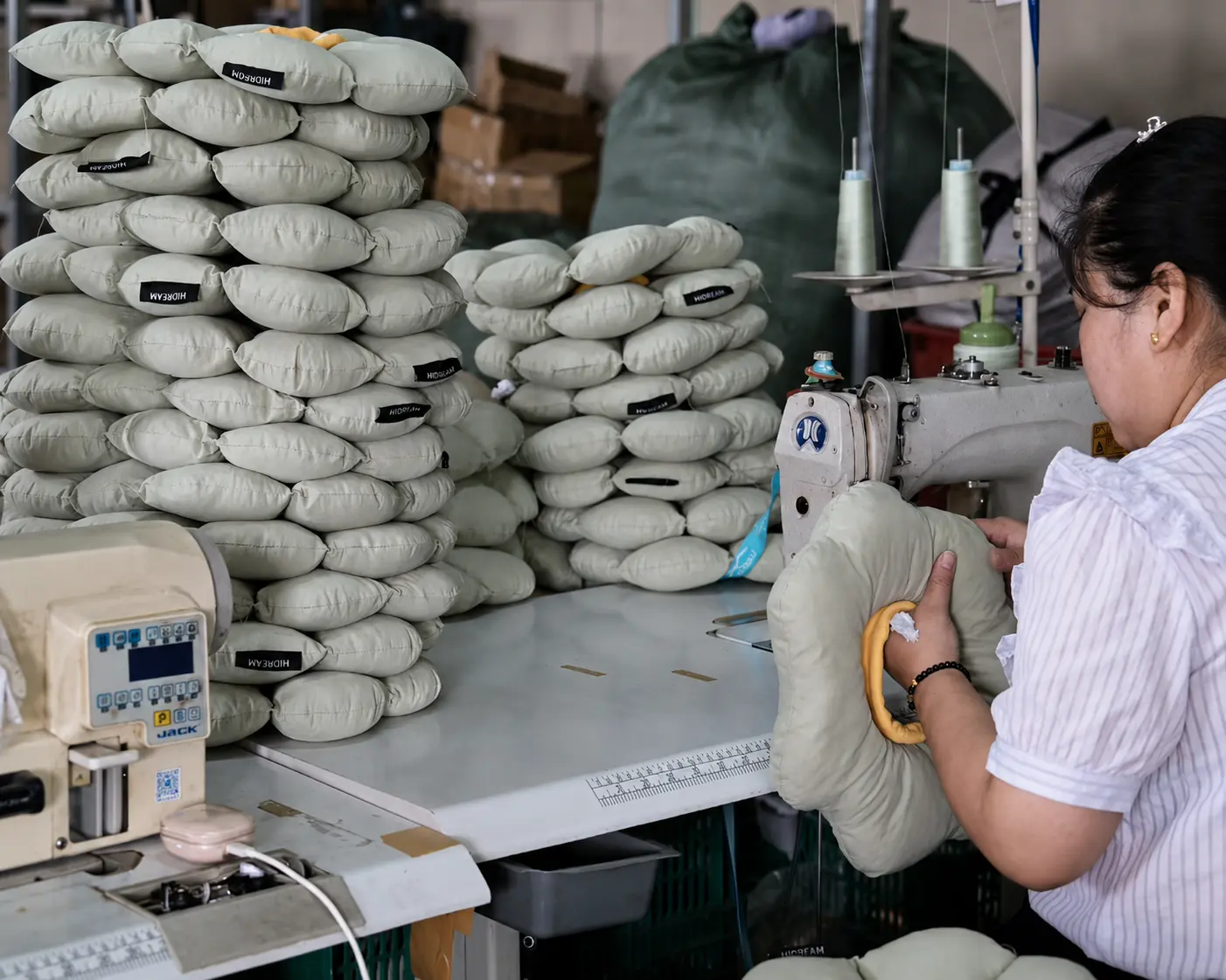 Worker at a sewing station beside stacks of pet bed cushions.